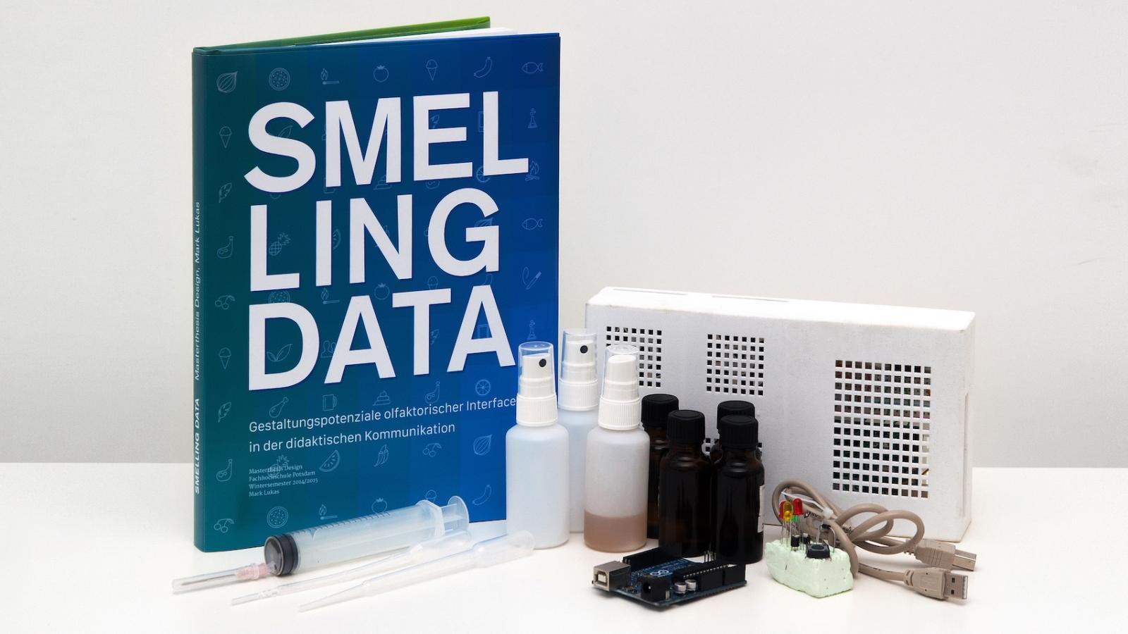 Aufgestelltes Buch mit der Aufschrift "Smelling Data". Daneben verschiedene Gegenstände wie kleine Sprühflaschen und USB-Kabel. 