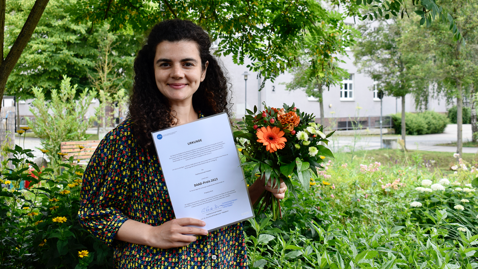 Die Preisträgerin des DAAD Presies, Phantina Sholi hält ihre Urkunde und ein Strauß Blumen hoch. Sie steht in einem grün bewachsenen Garten.
