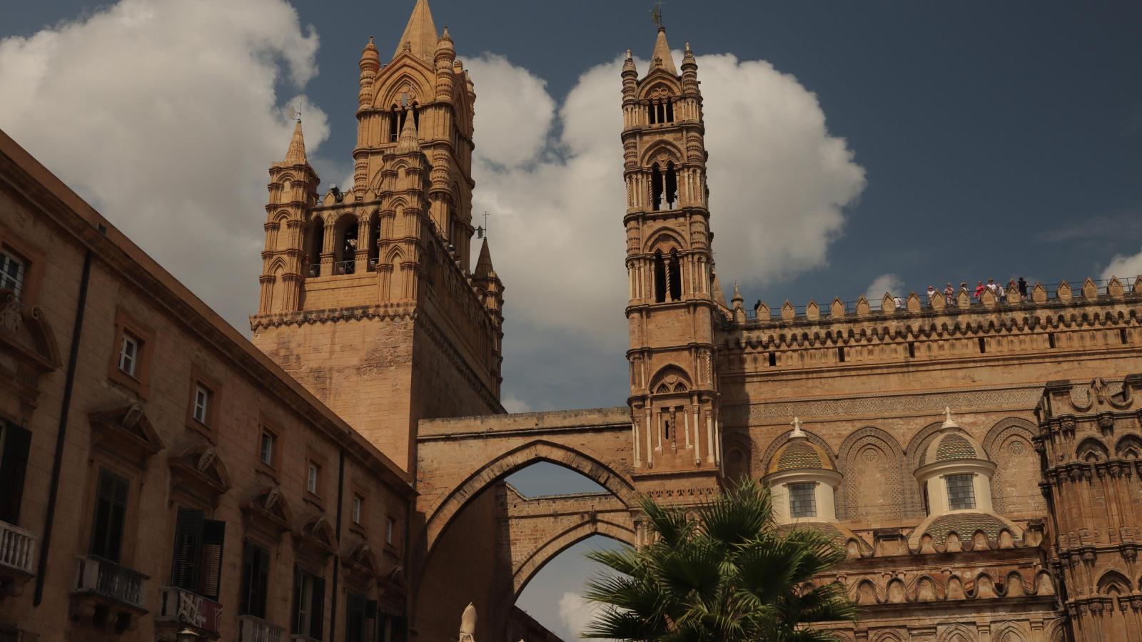 Vor einem blauen, leicht bewölkten Himmeln ist die große, alte Kathedrale von Palermo zu sehen. Sie ist aus hellem Stein. Zwei Türme strecken in die Höhe und sind mit einem Torbogen verbunden.