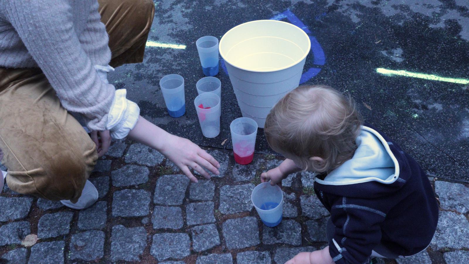 Kleinkind und Erwachsener schauen sich Plastikbecher mit Farbinhalt an