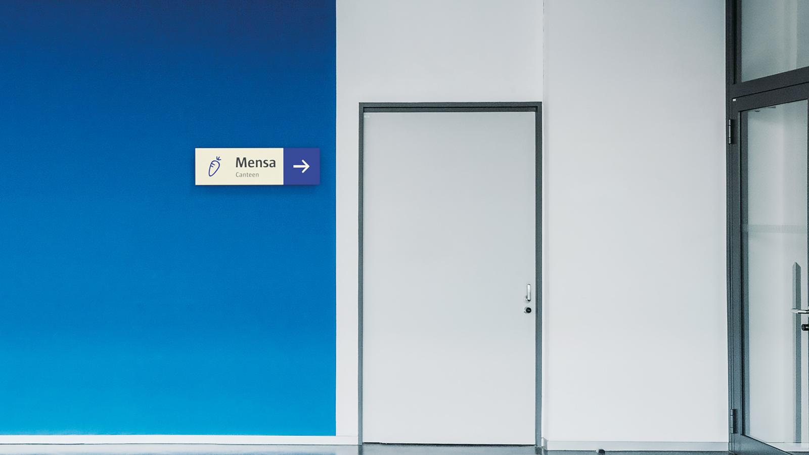 Blaue Wand vor dem Eingang zur Mensa mit Richtungspfeil und Auszeichnung "Mensa/Canteen"; daneben das Symbol einer Karotte