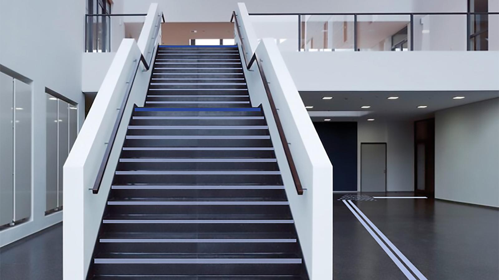 Foyer des Hauptgebäudes mit Treppe: Blindenleitstreifen führen von der Mensa zum Ein-/Ausgang. Vor der Treppe kleben Punkte auf dem Boden.