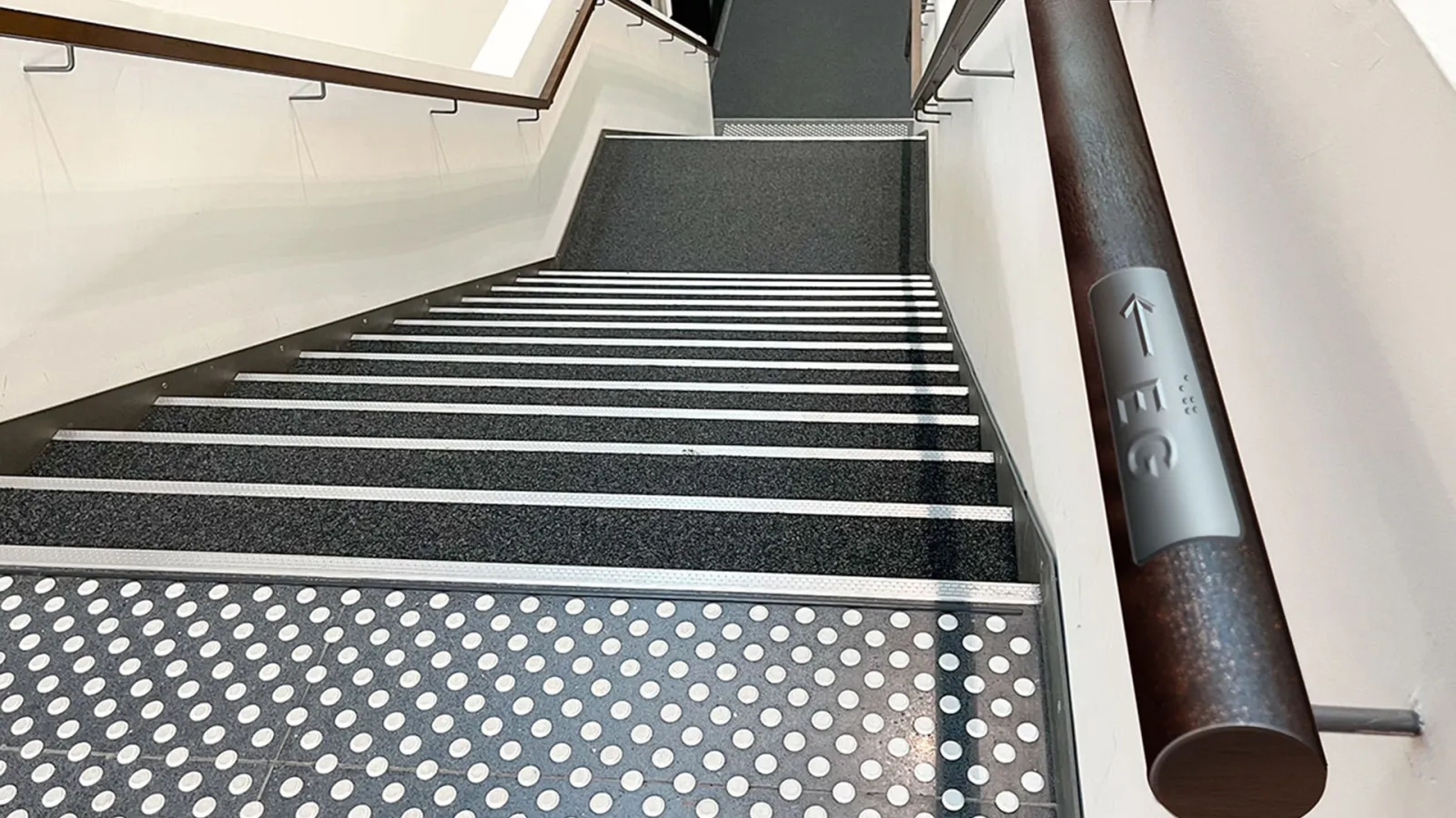 Treppe in der Bibliothek von oben; Auf dem Geländer befindet sich eine Plakette mit erhabener Schrift "EG" und Pfeil sowie Blindenschrift