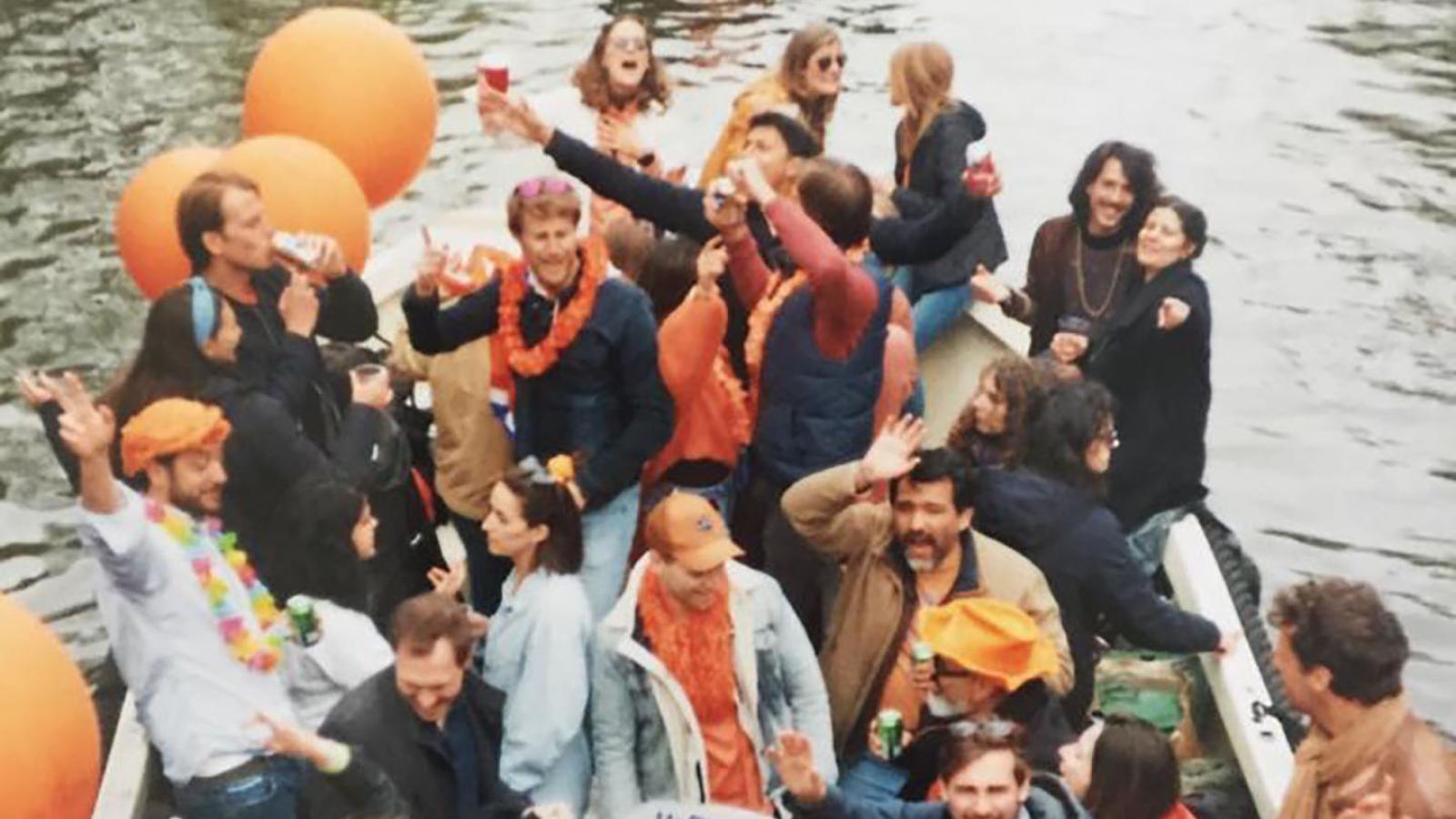 Im Vordergrund sind viele Menschen, die Meisten mit orangener Kleidung auf einem Bod zu sehen. Im Hintergrund sind andere kleine Boote auf dem Wasser.