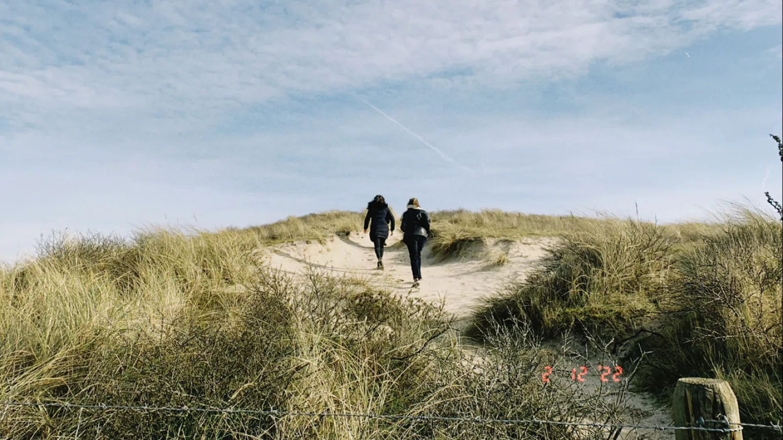 Zwei Personen laufen auf einem Sandweg eine Düne Hoch. Der blaue Himmeln ist leicht bewölkt. 