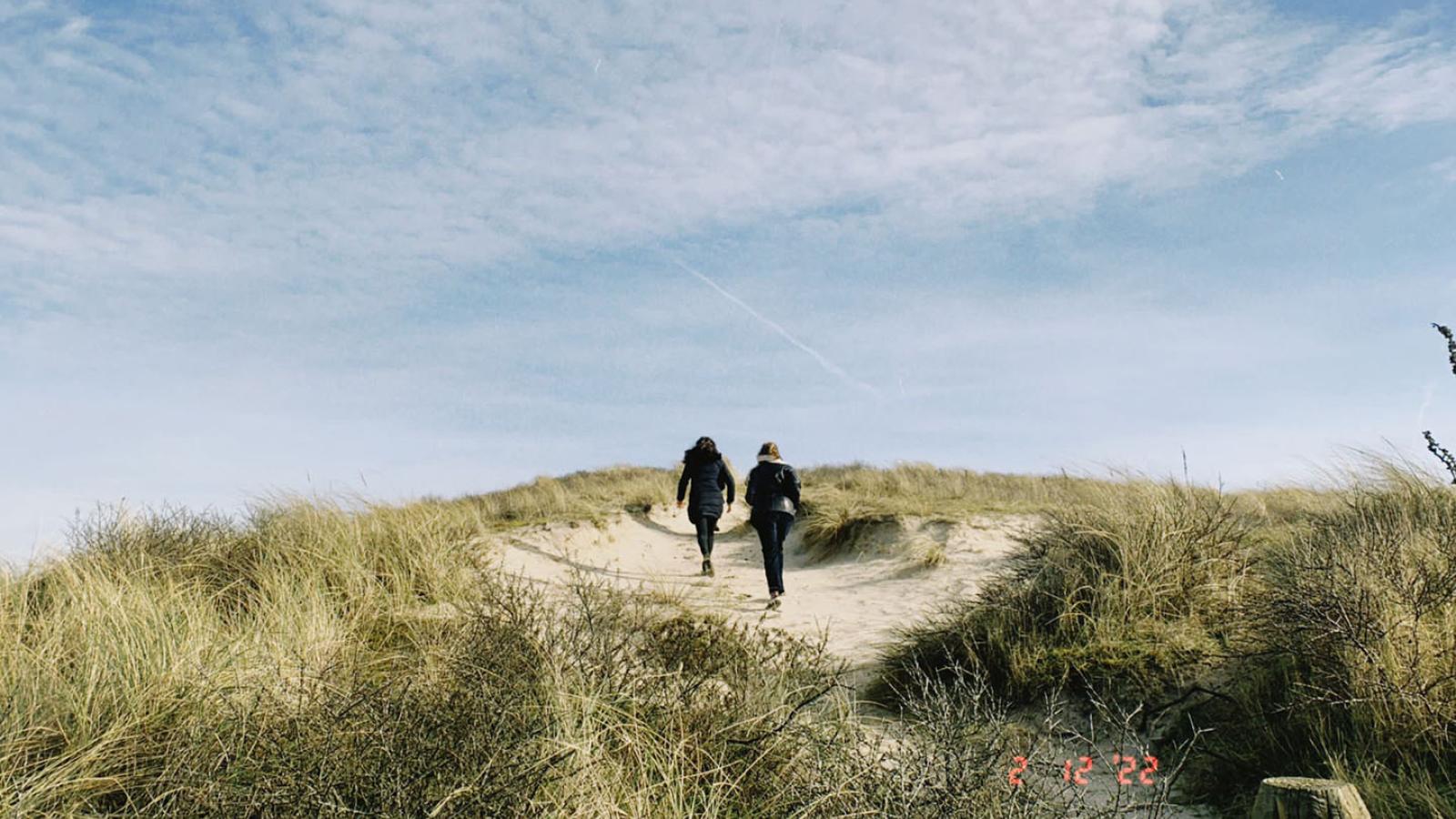 Zwei Personen laufen auf einem Sandweg eine Düne Hoch. Der blaue Himmeln ist leicht bewölkt. 