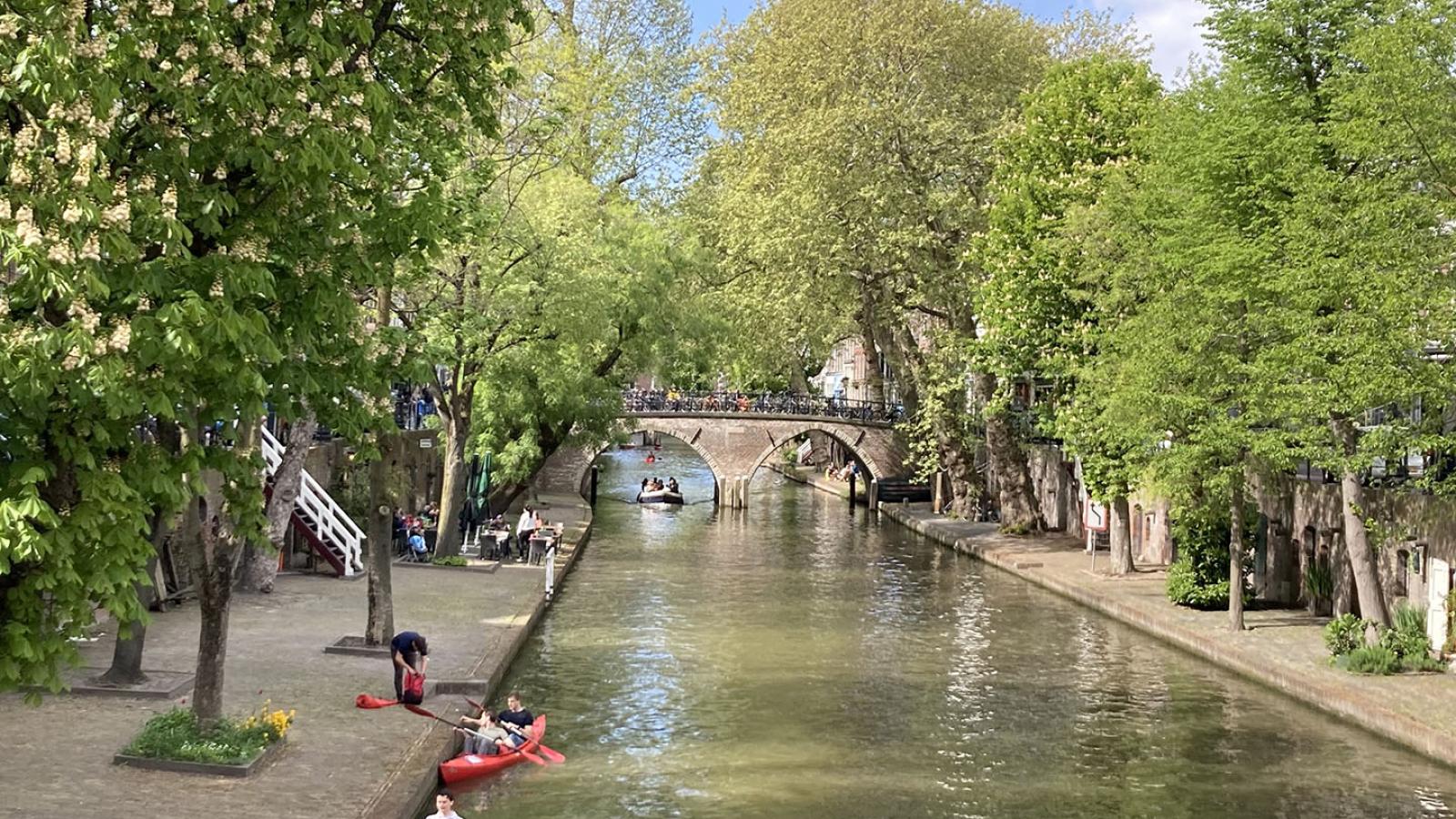 Man blickt auf einen stillen Kanal und eine kleine Brücke aus Stein die diesen überquert. Das Ufer ist mit Bäumen bepflanzt die viele Grüne Blätter tragen. Zwei Personen paddeln einzeln im Kanu.