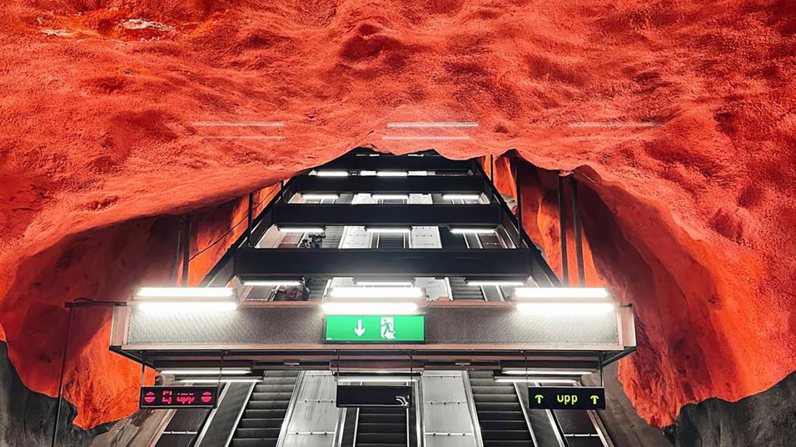 Drei U-Bahn-Rolltreppen führen weit nach oben. Die Rolltreppen sind von einer roten Masse umgeben die wie Kunststoff oder Stein aussieht.