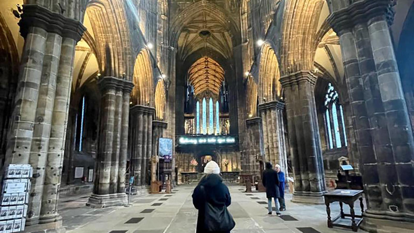 Ein Blick von innen in die Glasgower Kathedrale St. Mungo's Catherdral. Eine Person steht mittig mit dem Rücken zur Kamera. Der Raum ist Dunkel aber die Torbogen sind von unten beleuchtet.