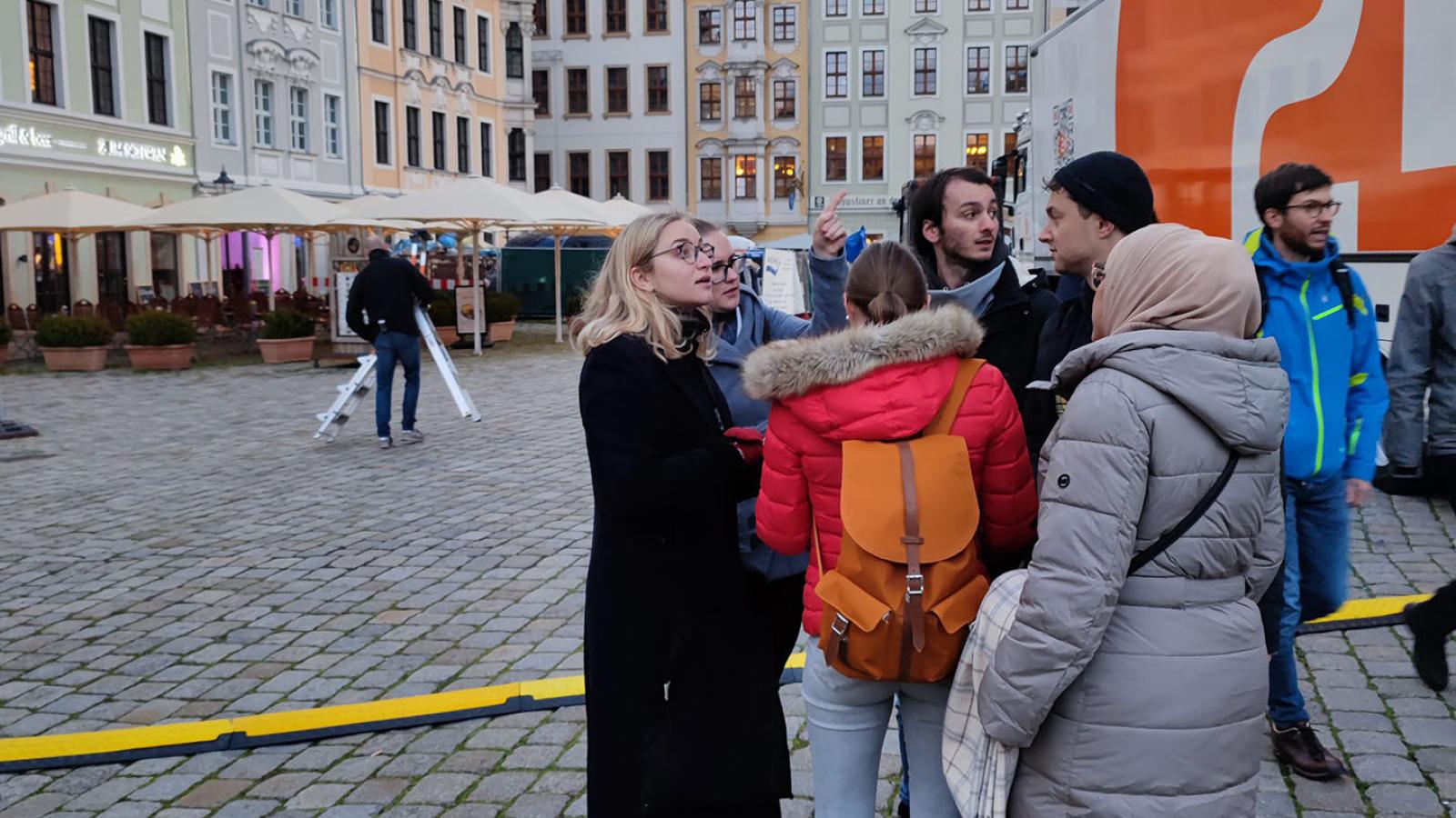 Eine kleine Gruppe Menschen in Winterklamotten stehen auf einem Platz in Dresden mit dem Rücken zur Kamera. Der Himmel ist grau. 
