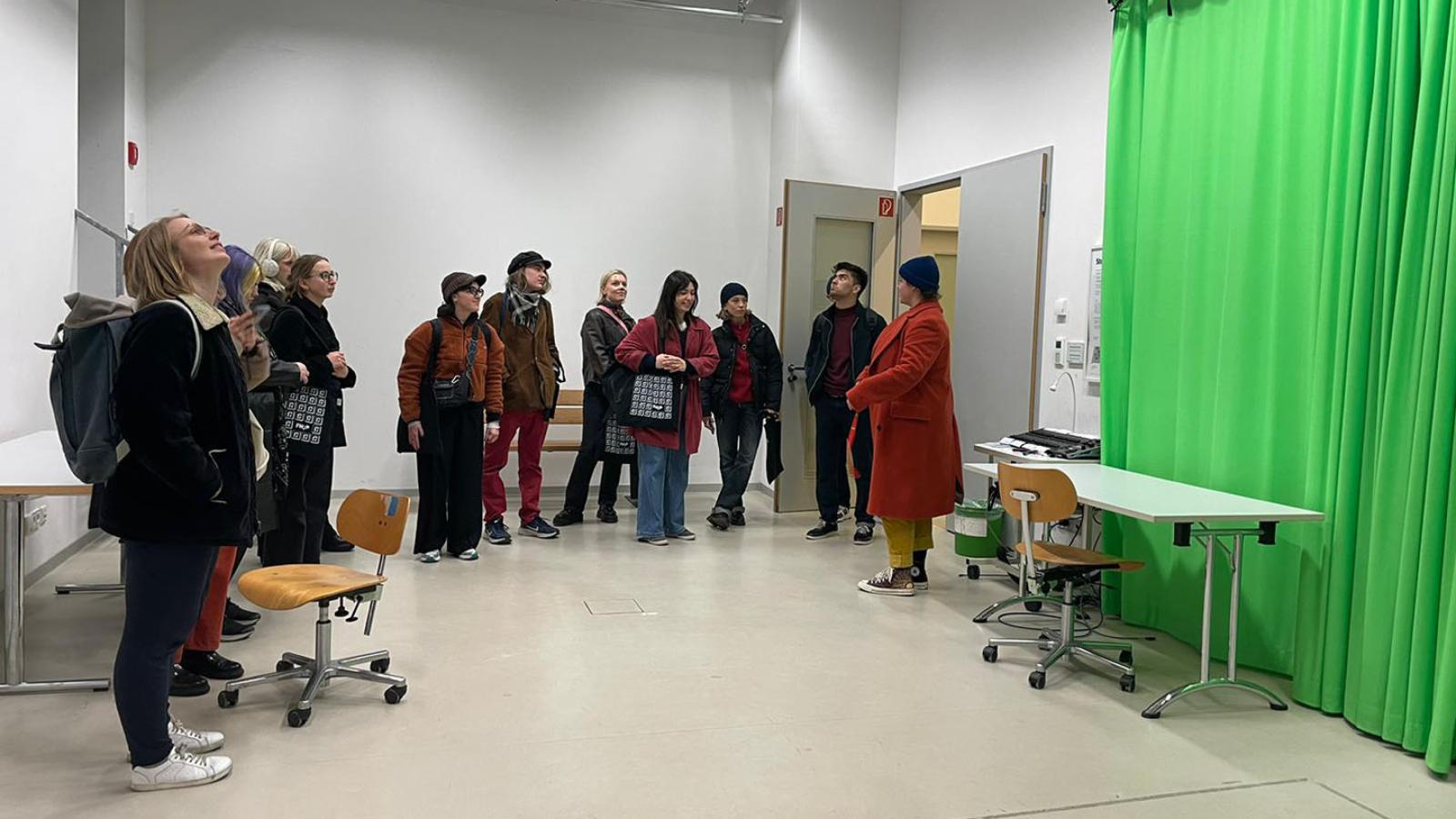 Eine Gruppe internationaler Studierende steht am linken Ran eines Raumes. An der rechten Wand hängt ein knall-grüner Vorhang.