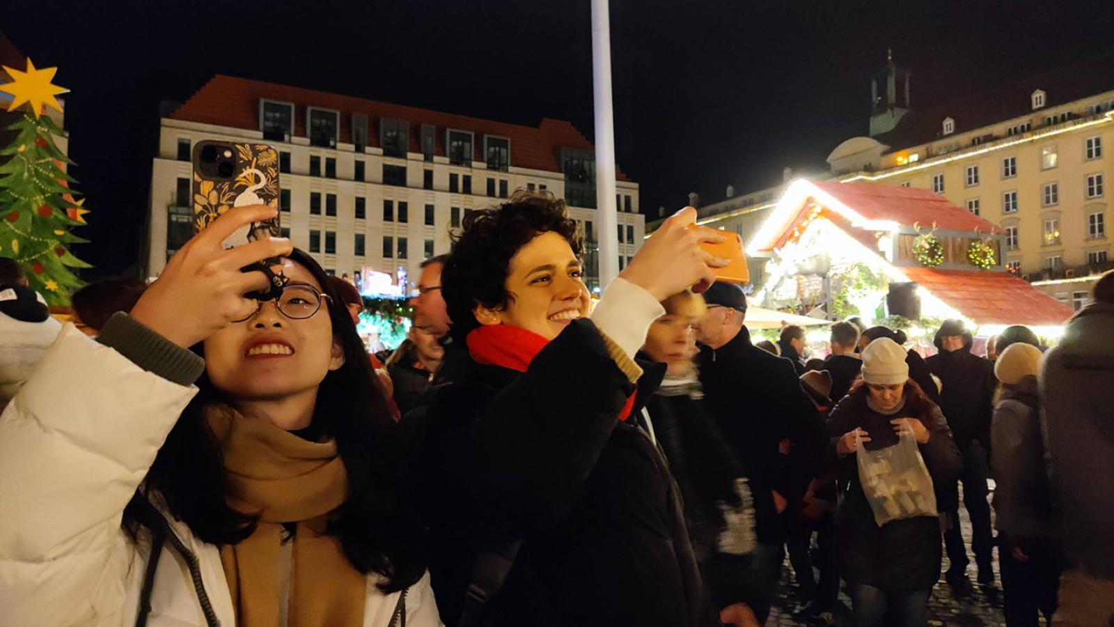 Zwei Personen stehen in der unteren linken Ecke des Bildes und halten ihre Handys in die Höhe als würden sie fotografieren. Sie stehen auf einem Weihnachtsmarkt. Der Himmel ist dunkel aber die Lichter vom Markt glänzen hell.
