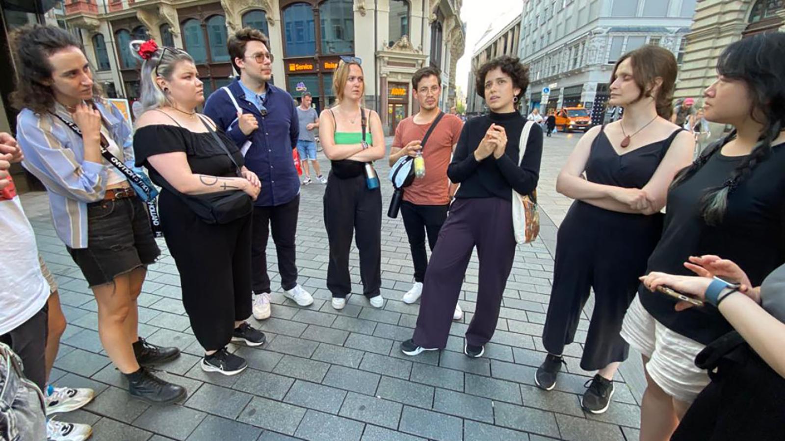 Ungefähr zehn Menschen stehen in einem Halbkreis auf einem Platz in Leipzig und schauen auf eine Person die Teil des Kreises ist und etwas erzählt.