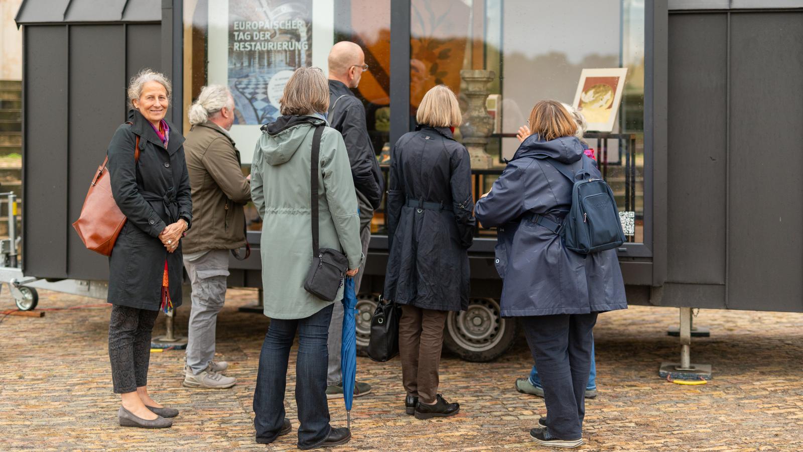 Mitarbeiter*innen der SPSG und der FHP betrachten die Ausstellung der Restaurierung der FHP durch das Schaufenster des FHP Mobils vor dem Neuen Palais in Potsdam