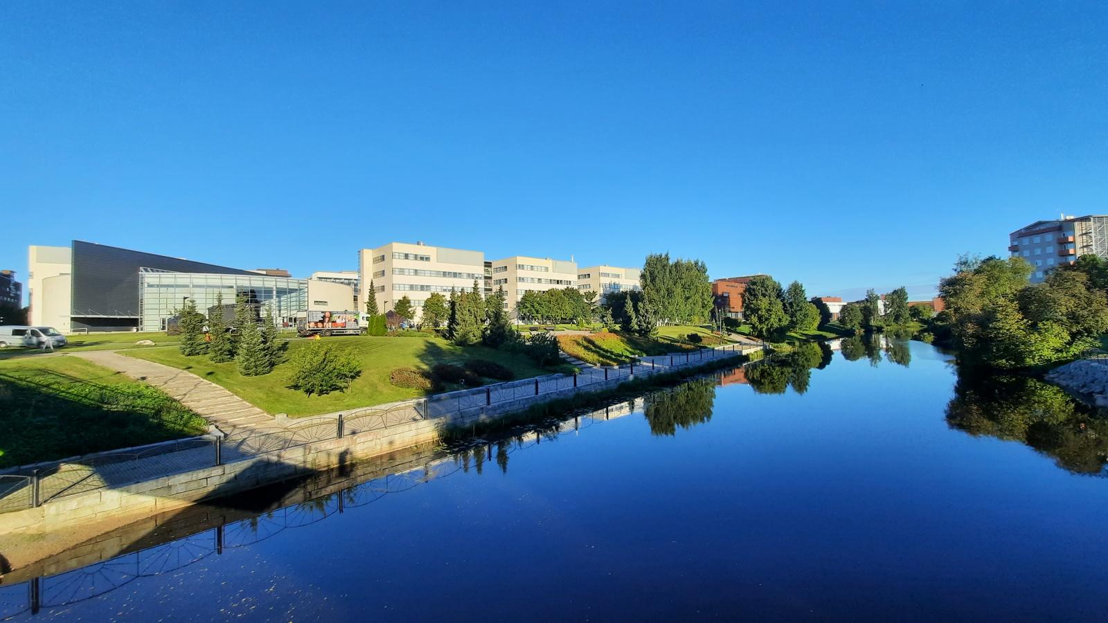 Die modernen, eckigen Gebäude der Seinäjoki University of Applied Sciences sind unter einem wolkenlosen blauen Himmel an einem Flussufer zu sehen.