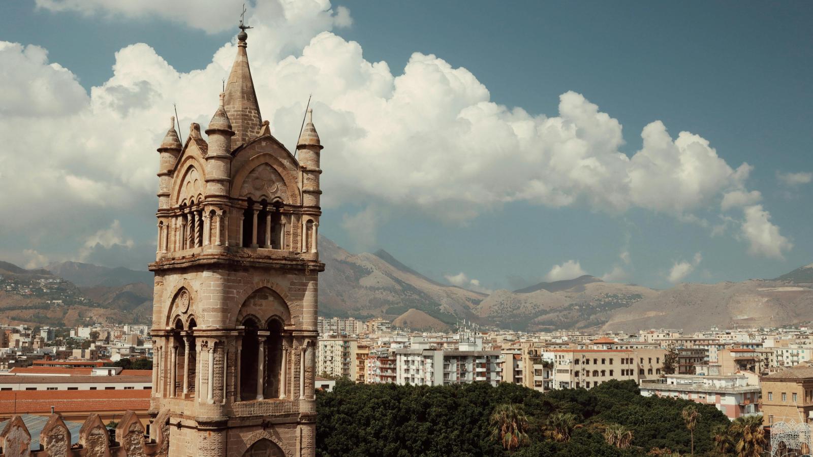 Im Vordergrund auf der linken Seite des Bildes ist ein alter Kirchturm zu sehen - er ist aus hellem Stein. Im Hintergrund ist ein weiter Blick über die Dächer von Palermo. Hinter den Gebäuden ragen Berge in die Höhe.