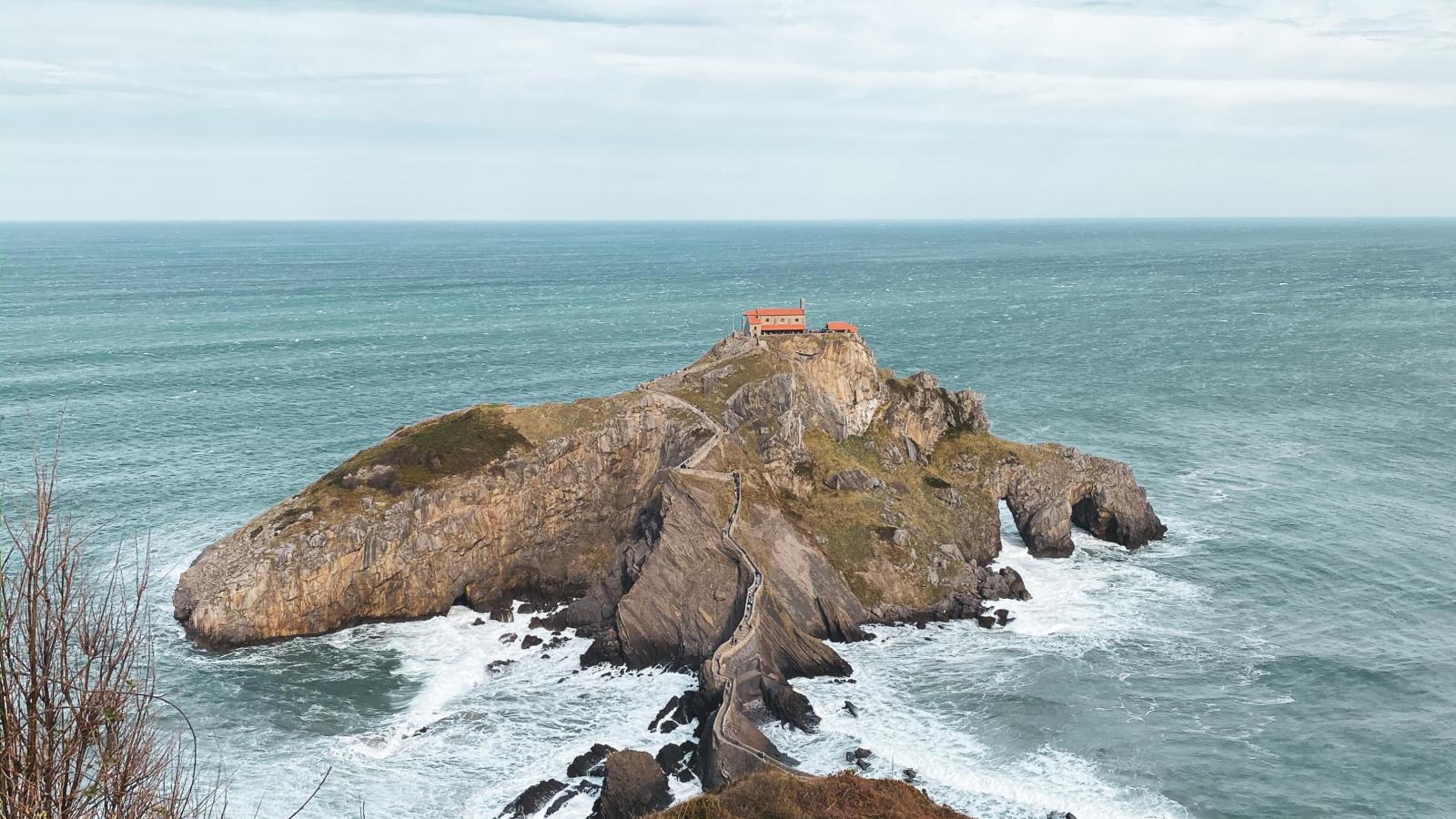 Von einer Klippe wird aufs Meer rausgeschaut. Es streckt sich ein dünner Weg über Stein zu einer kleinen Stein-Insel heraus auf dessen Spitze zwei Häuser sind.