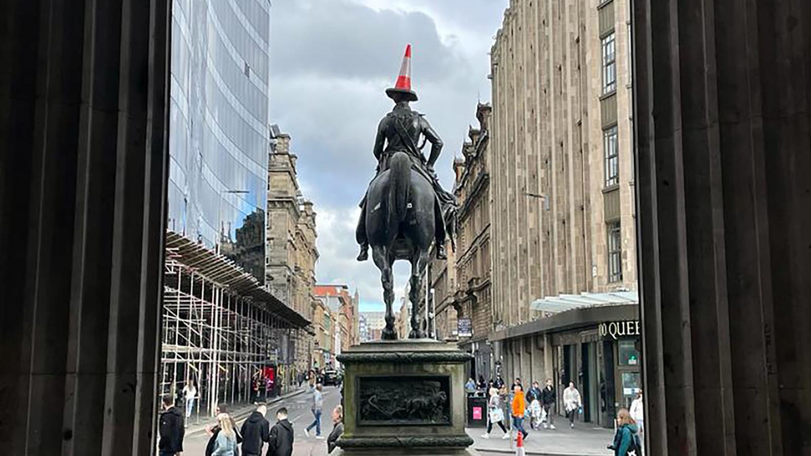 Der Blick führt aus dem Eingang des Gallery of Modern Art Glasgow heraus auf eine Statue auf einem Pferd die man von hinten sieht. Auf dem Kopf der Figur ist ein Verkehrskegel balanciert.