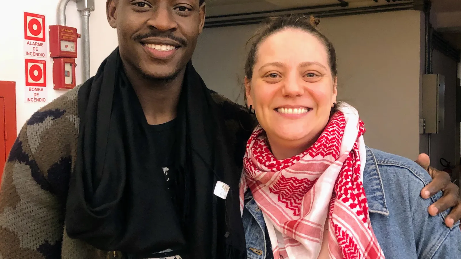 Steve Seide, Migrant aus Haiti, trägt schwarzen Schal und gemusterten Pullover, steht neben Marina Volpatto mit rotem und weißem Schal und Jeansjacke, aufgenommen im Rahmen des Podcasts 'Entre Caminhos' zur sozialen Arbeit mit Migrant*innen in Brasilien
