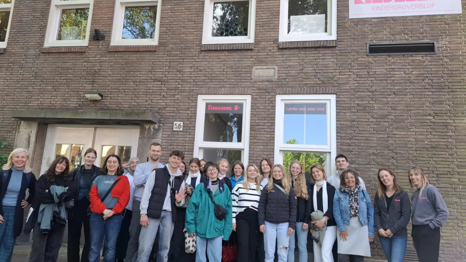 Eine Gruppe Studierender steht vor dem Kindergarten Mauritsgade, erkennbar am Schild mit dem Schriftzug 'Kinderrijk Kinderdagverblijf' an der Backsteinfassade.