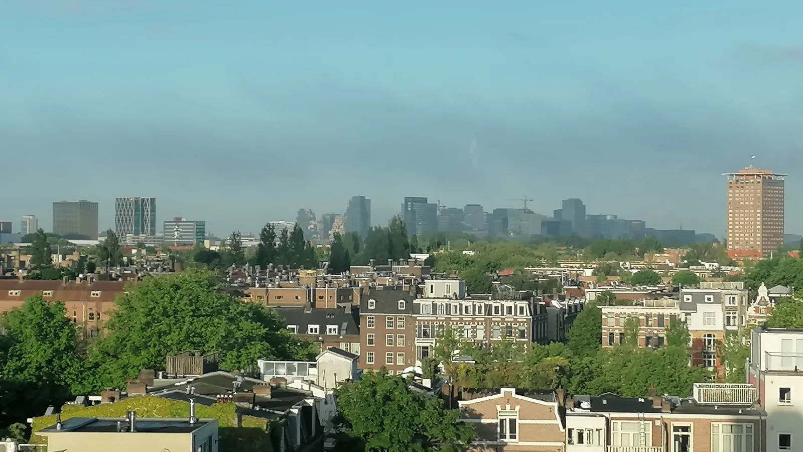 Stadtpanorama von Amsterdam vom Volkshotel nahe der Universität aus mit Wohnhäusern im Vordergrund und Hochhäusern im Hintergrund unter blauem Himmel mit leichter Bewölkung.