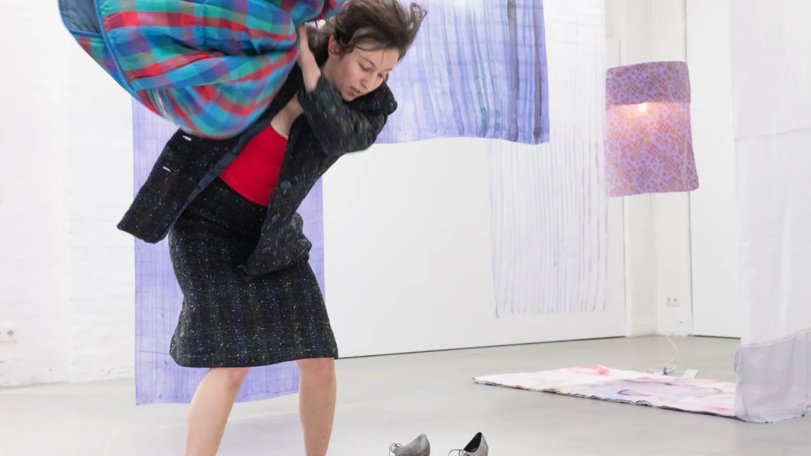 Das Bild zeigt eine Person, barfuß, in einer Ausstellung mit Textilien, die sich eine karierte Decke über die Schultern wirft.. 