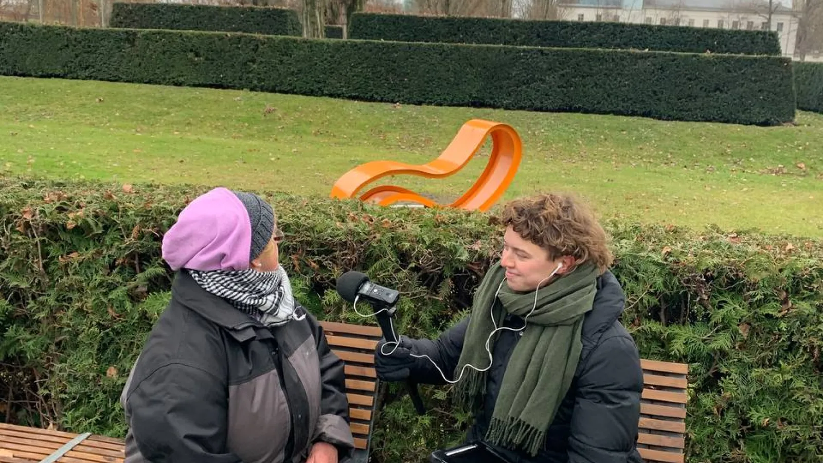 Das Bild zeigt zwei Personen auf einer Bank sitzend mit einer Parkanlage im Hintergrund.