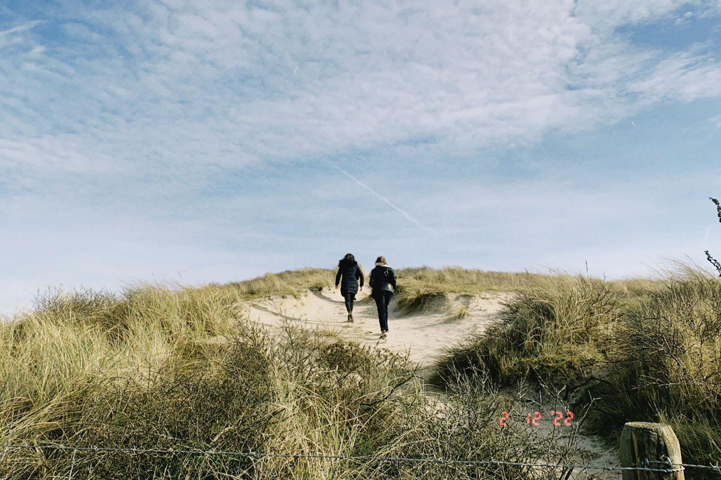 Zwei Personen laufen auf einem Sandweg eine Düne Hoch. Der blaue Himmeln ist leicht bewölkt. 