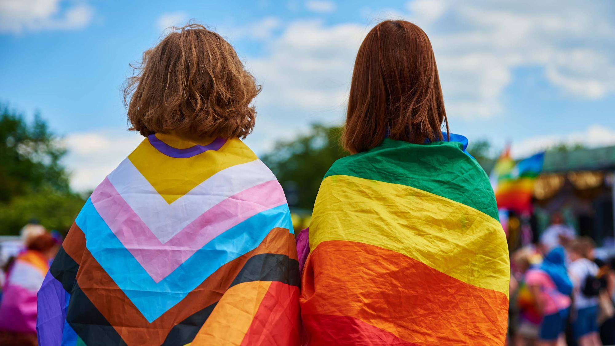 Zwei Personen, von hinten fotografiert, mit Regenbogen-Flaggen auf den Schultern