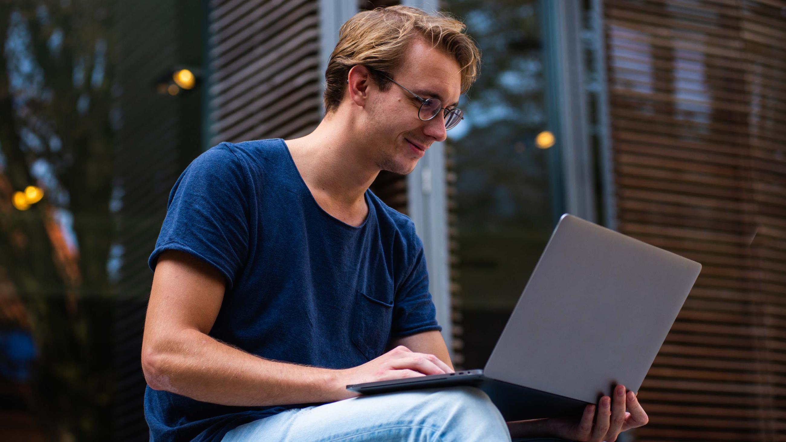 Ein fröhlicher Student sitzt vor einem Gebäude mit Laptop auf dem Schoß