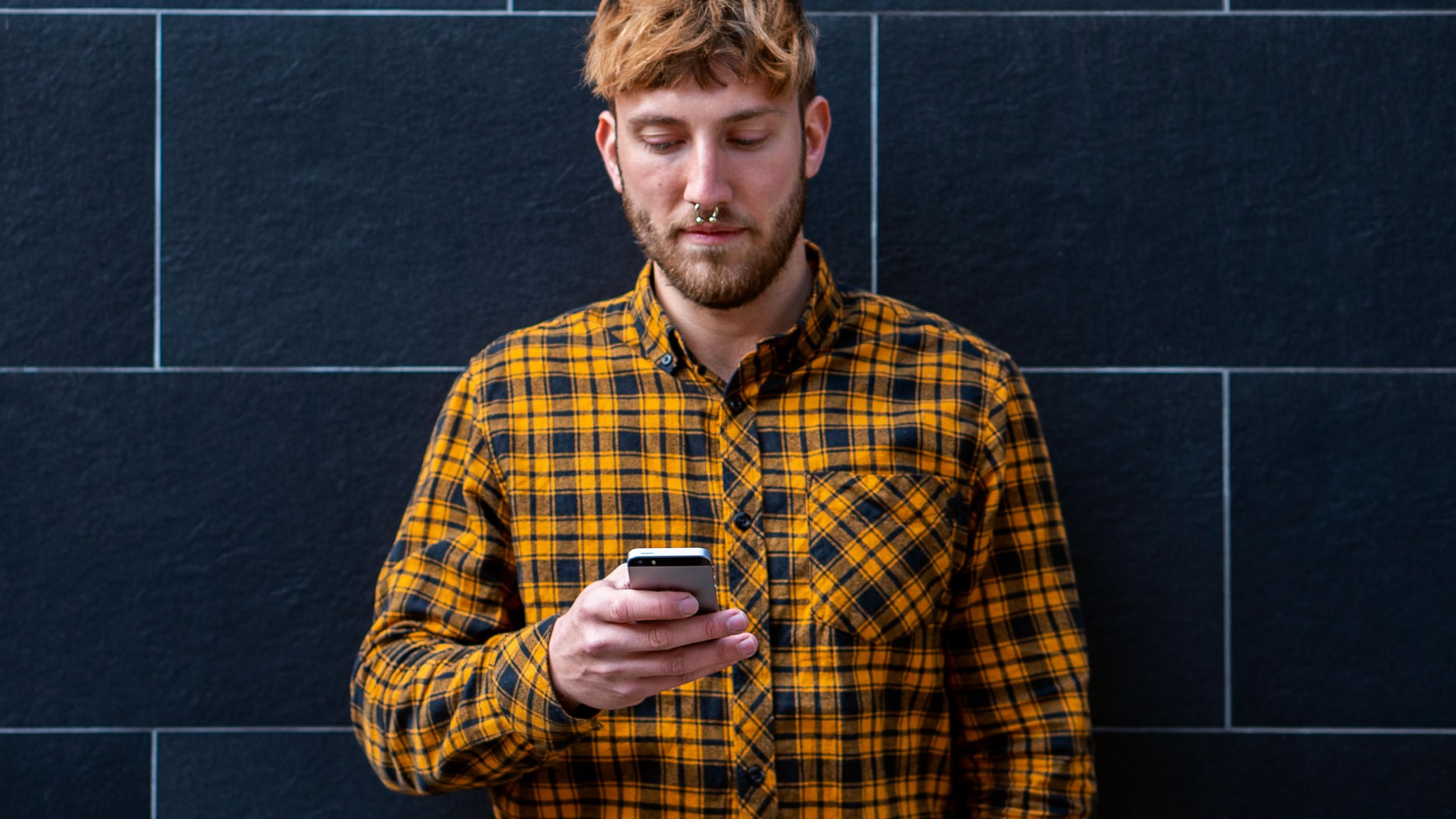 Ein Student im Karohemd steht vor einer dunklen Mauer und schaut auf ein Smartphone in seiner Hand.