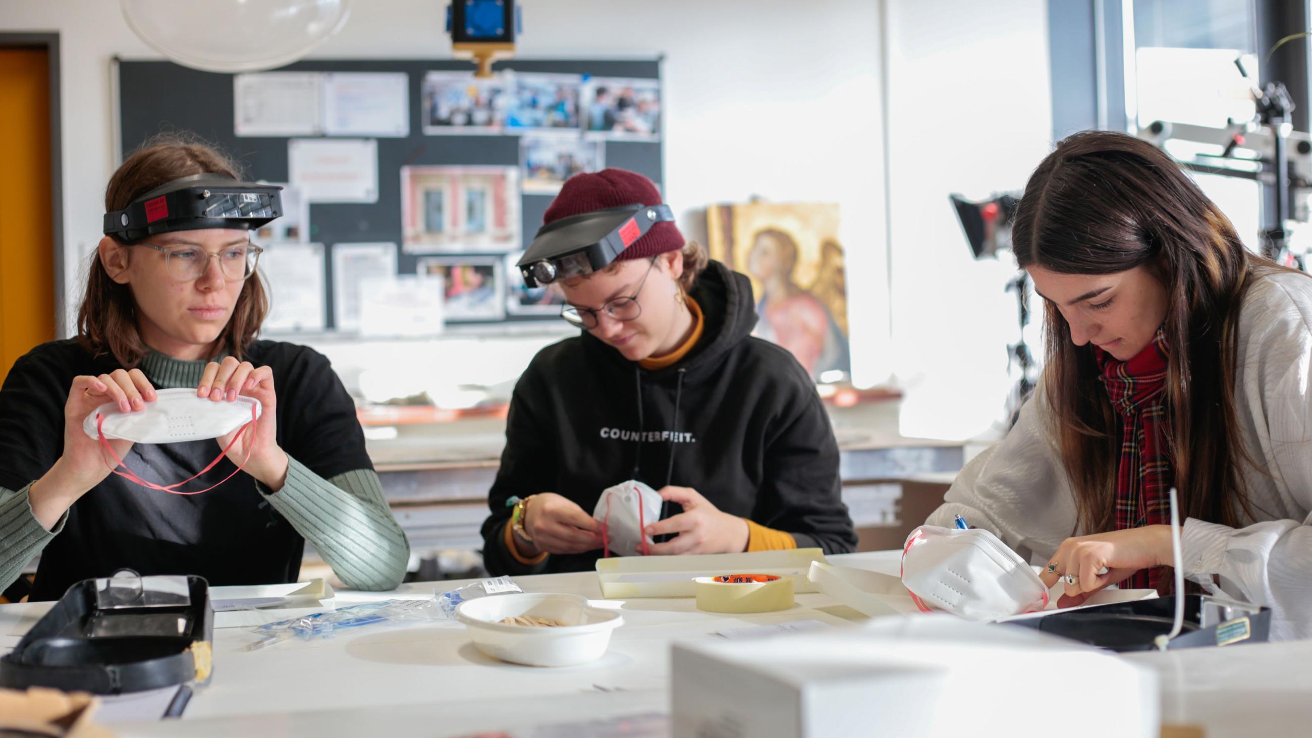 Drei Studierende, mit Schutzbrillen auf dem Kopf, arbeiten an einem Tisch.