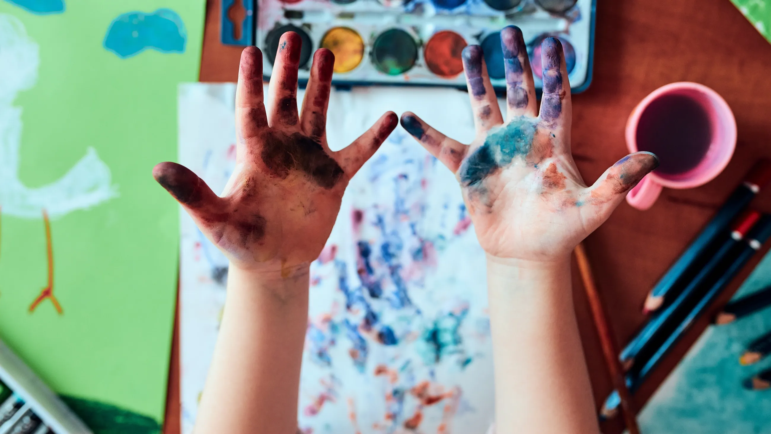 zwei bemalte Kinderhände vor Tischplatte mit Aquarellfarben, Malpapier und Pinseln
