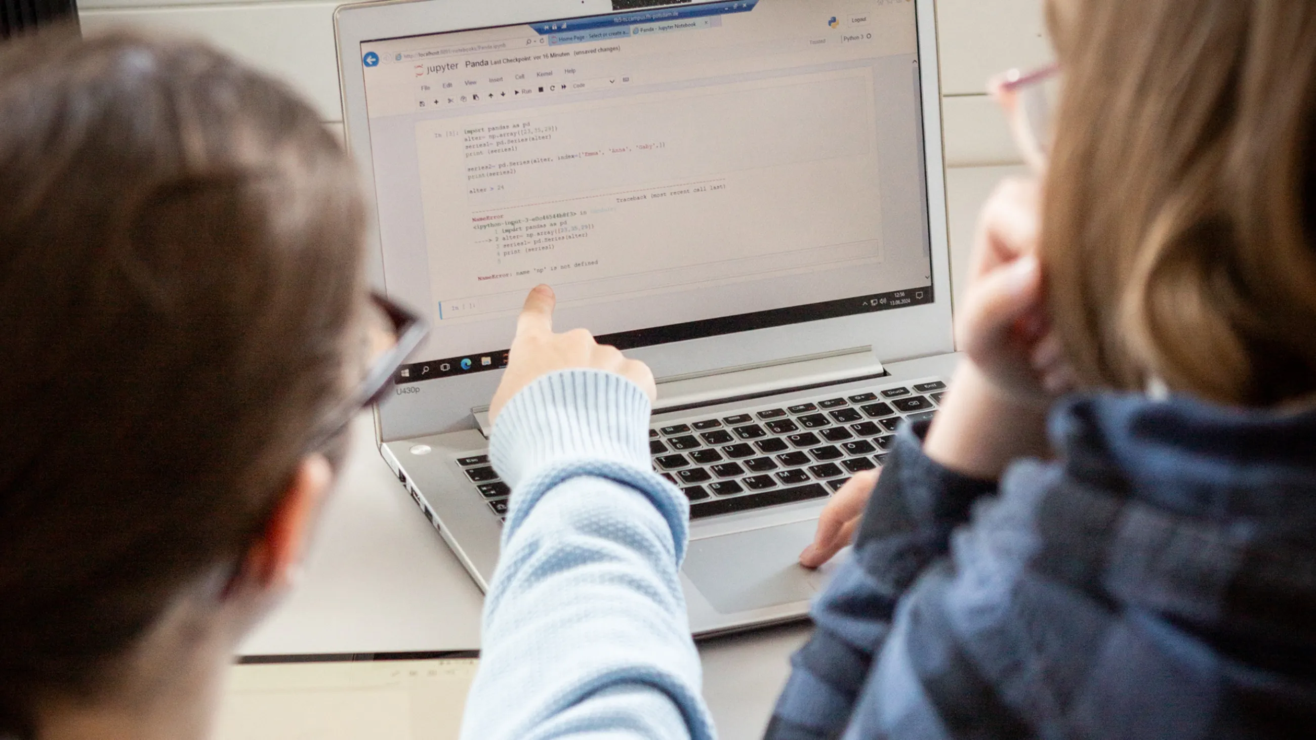 Zwei Studierende sitzen vor einem Laptop auf dem HTML-Codes zu sehen sind. Eine Person zeigt auf den Bildschirm.
