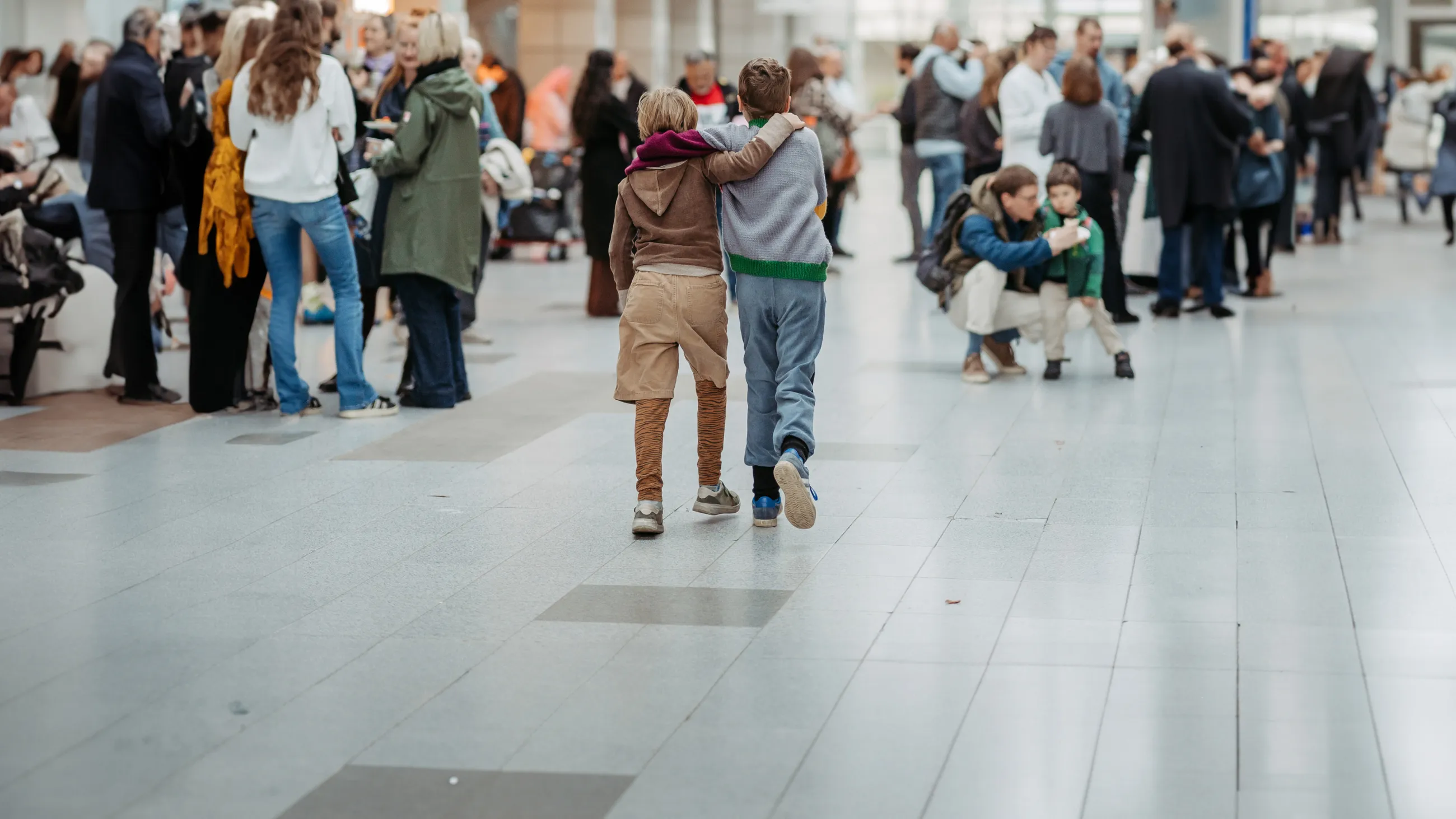 Zwei Kinder gehen Arm in Arm durch eine belebte Halle mit mehreren Menschen im Hintergrund.