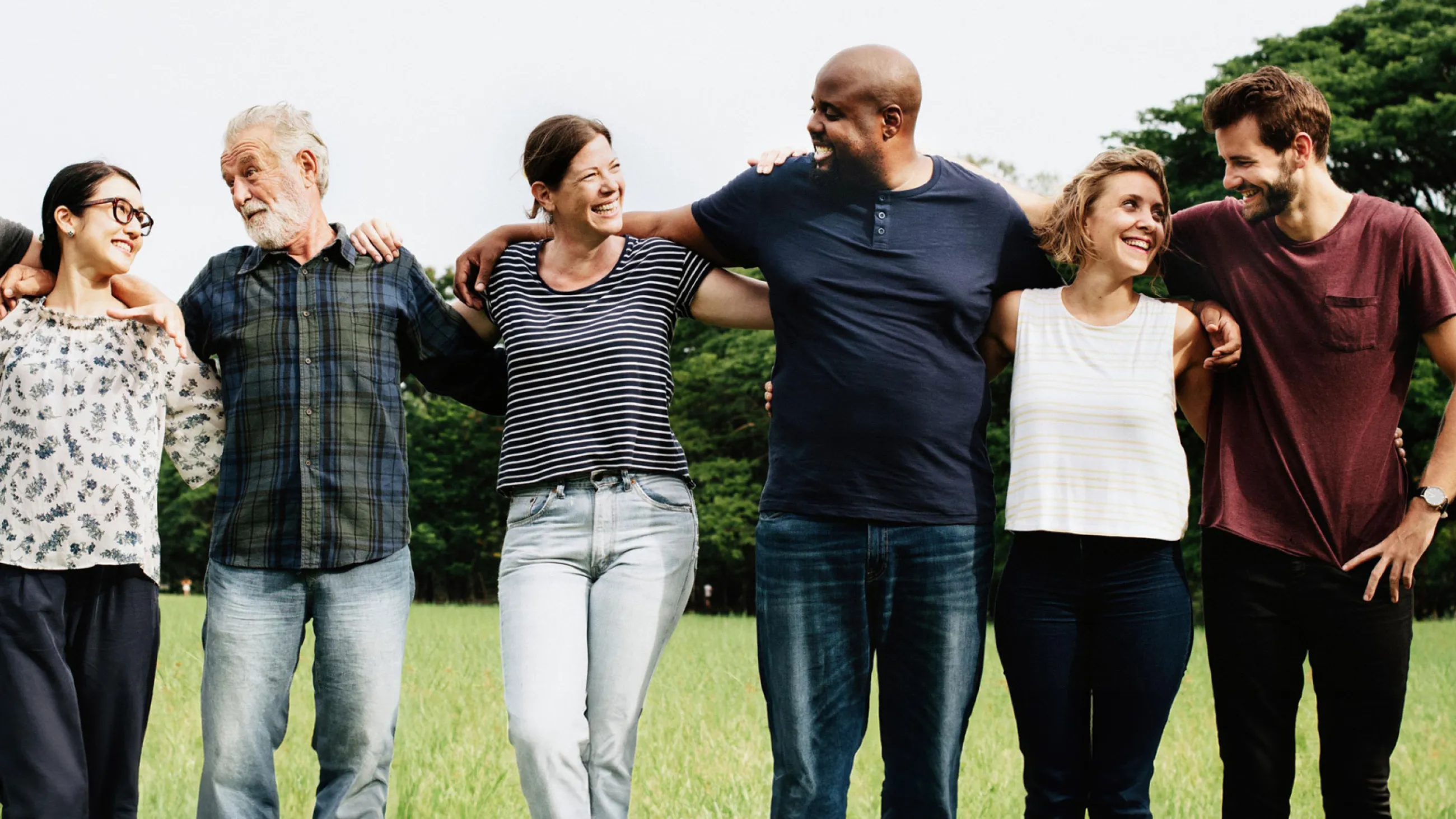 Sechs Menschen stehen eng nebeneinander auf einer Wiese und umarmen sich an den Schultern.