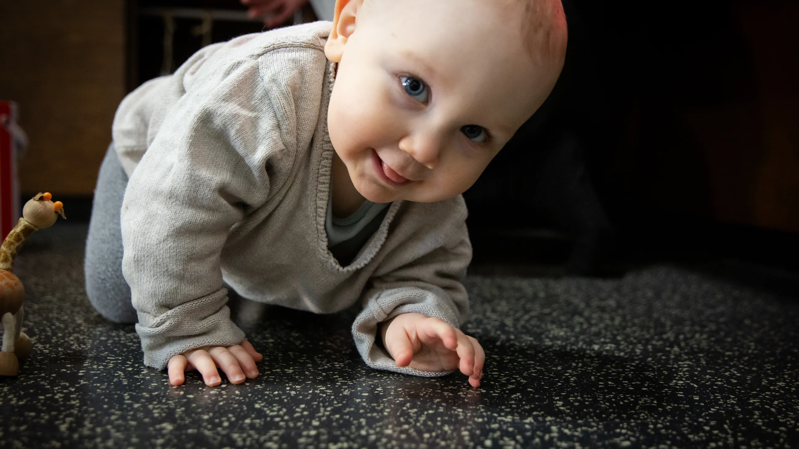 Kleinkind krabbelt auf einem dunklen, strukturierten Boden in einer Spielecke mit einem kleinen Spielzeugtier in der Nähe.