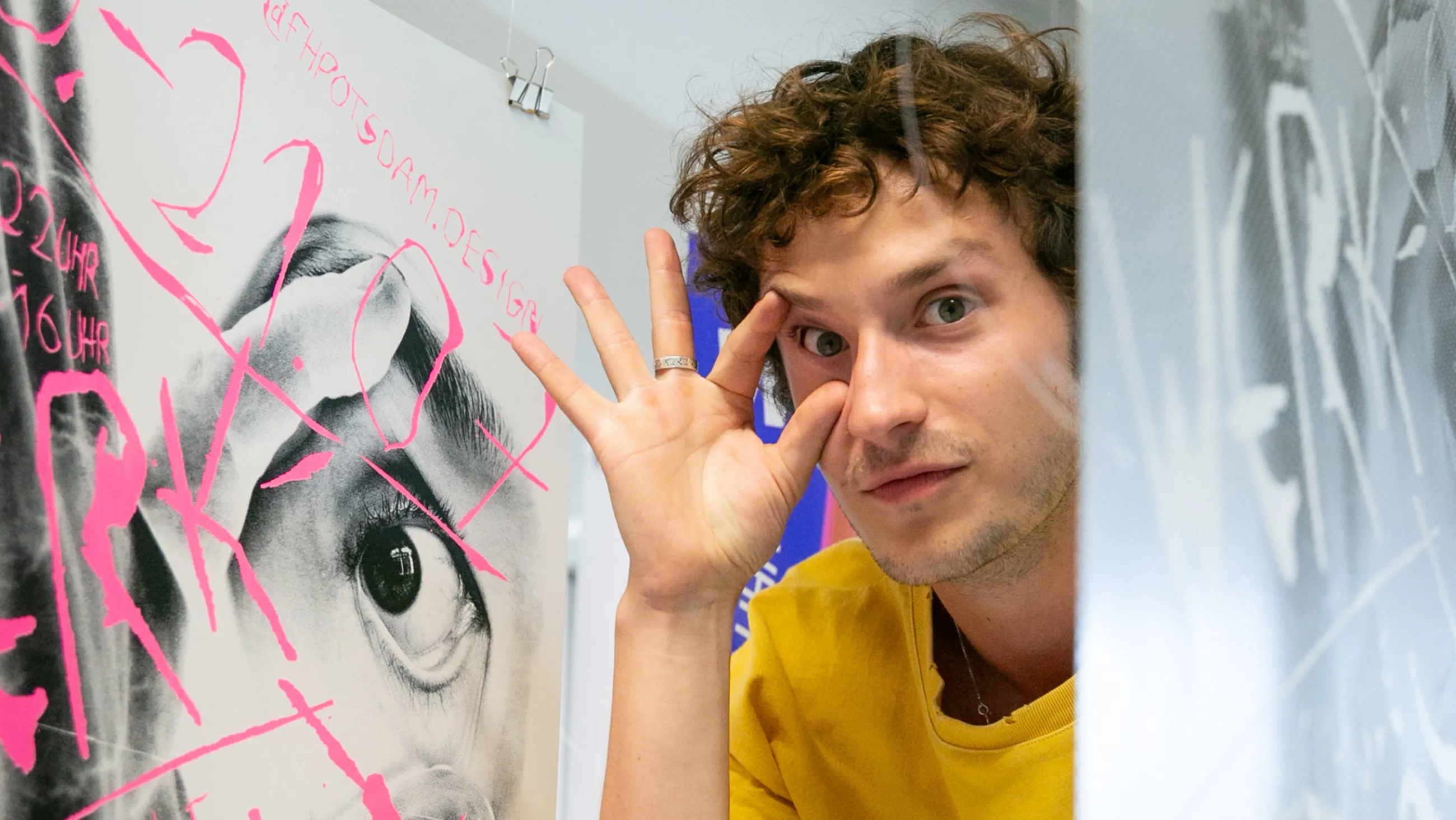 Ein Studierender posiert mit von Fingern aufgehaltenem Auge neben einem Bild einer Person, die sich das Auge mit Fingern offen hält