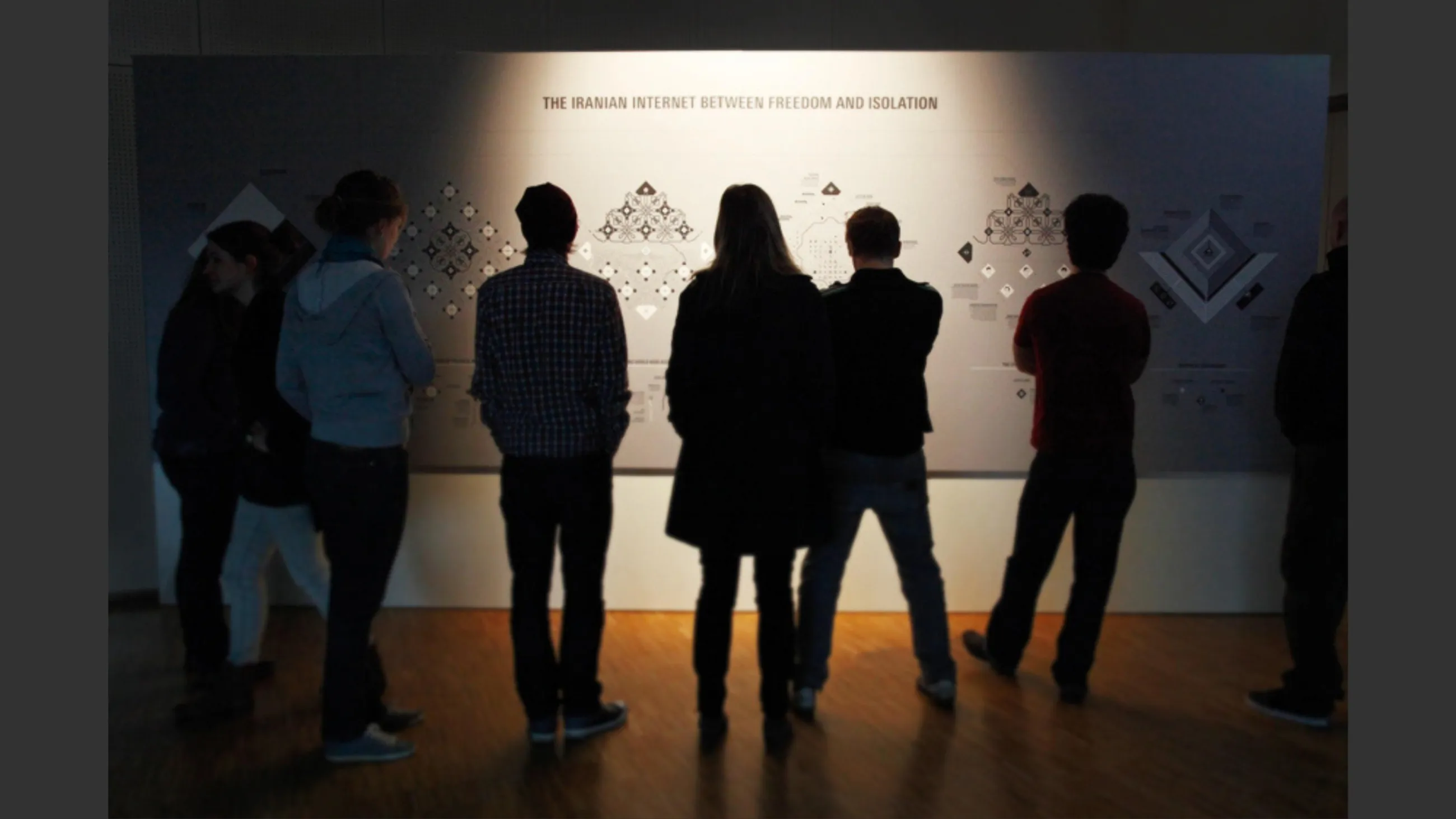 Mehrere Personen stehen in einem abgedunkelten Raum un betrachten eine Ausstellungswand zum Thema "The Iranian Internet"