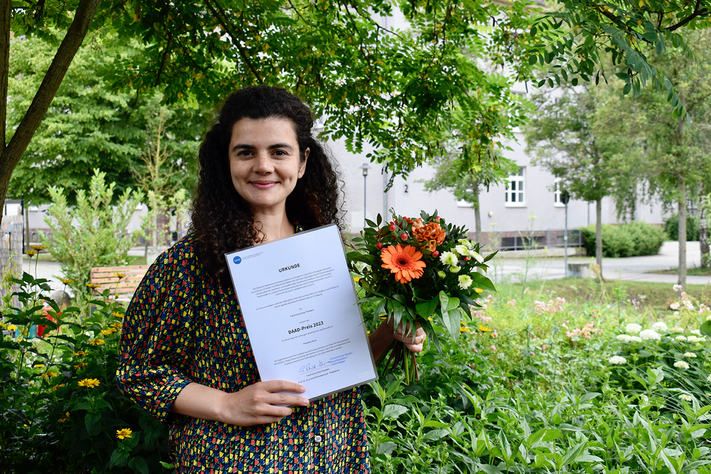 Die Preisträgerin des DAAD Presies, Phantina Sholi hält ihre Urkunde und ein Strauß Blumen hoch. Sie steht in einem grün bewachsenen Garten.