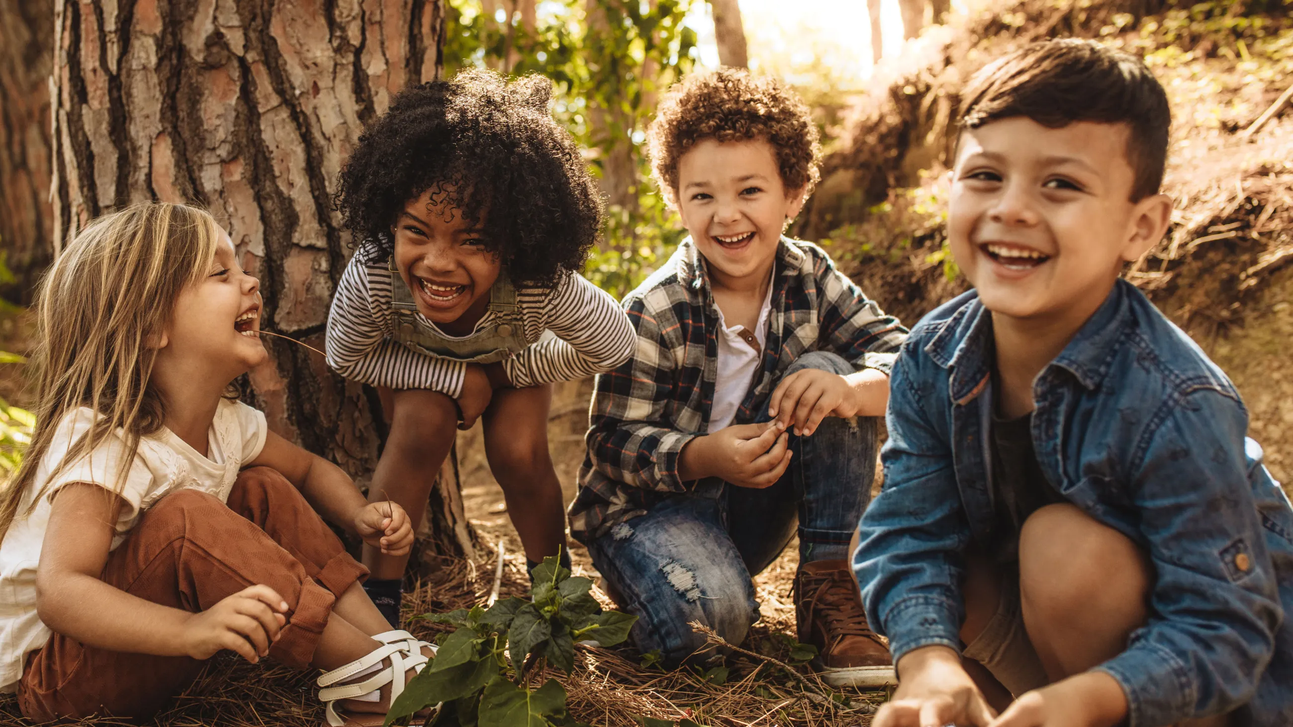 Vier lachende Kinder sitzen im Freien auf dem Boden vor einem Baum und schauen in die Kamera