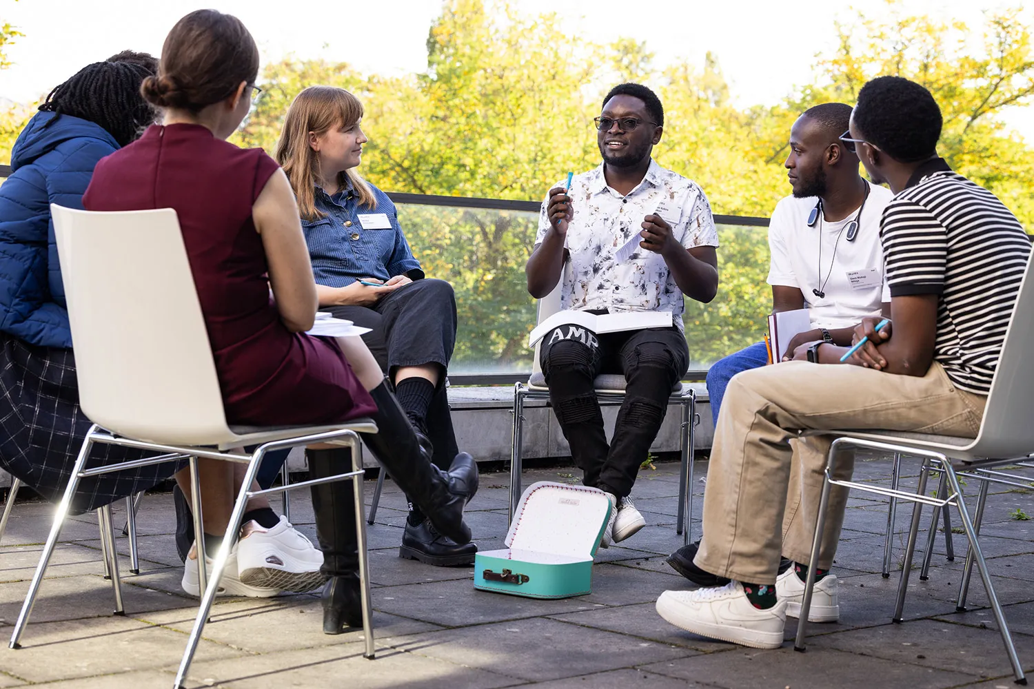 Sechs Personen sitzen im Kreis auf einer Terrasse vor herbstlichen Bäumen. Ein junger Mann mit Brille spricht und gestikuliert, die anderen hören ihm zu.
