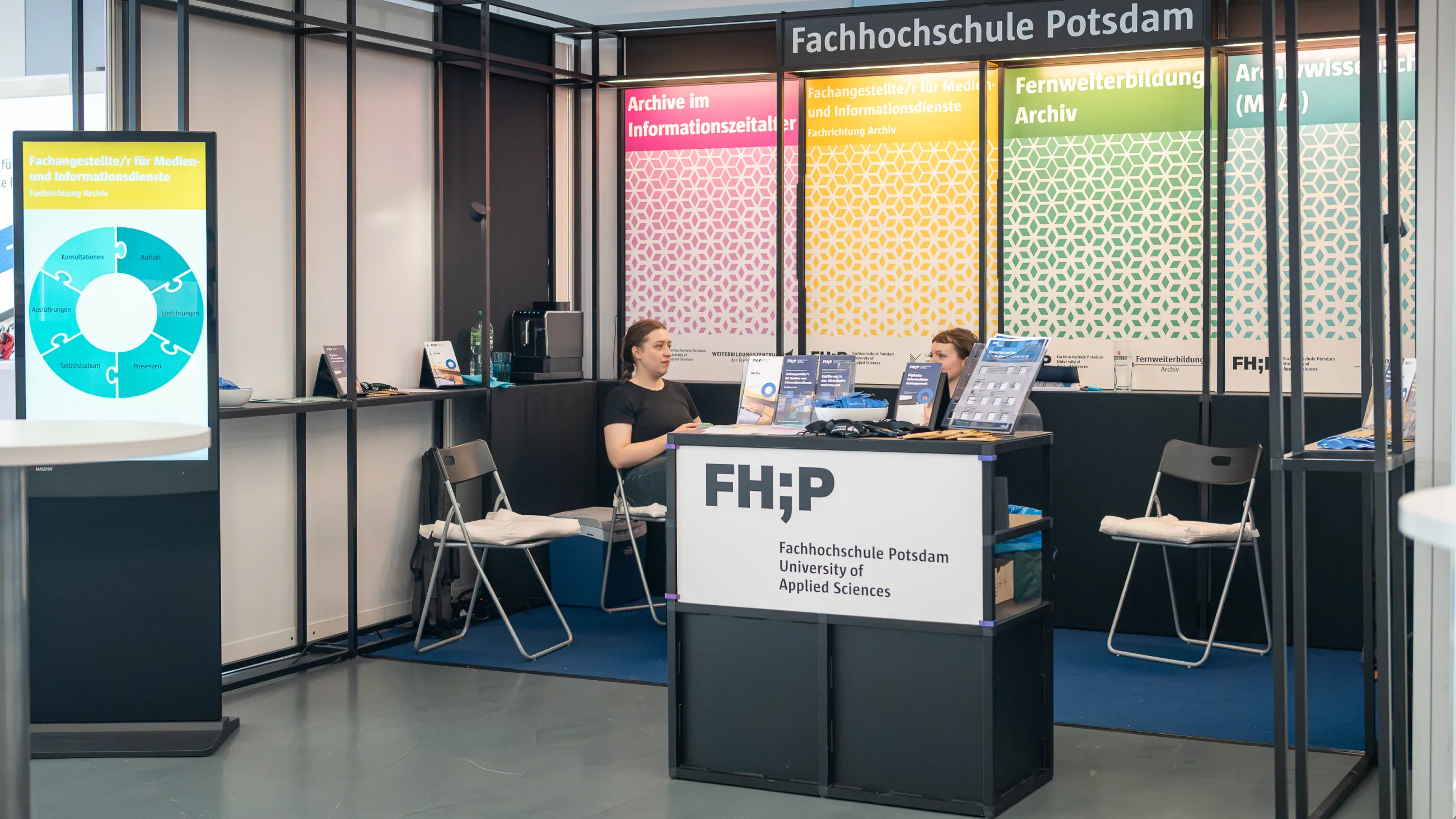 Stand der FHP auf dem Archivtag in Hof 2026. Zwei Frauen sitzen auf Klappstühlen und sprechen miteinander. Im Hintergrund sind Plakate mit dem Angebot der Fernweiterbildung Archiv.