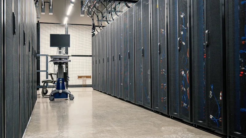 Dunkler Gang eines Serverraums. Etwas weiter hinten im Gang steht ein mobiler Tisch mit Computerterminal