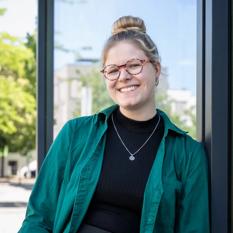 Laura Mareen Roesler, Studentin des Masterstudiengangs Frühkindliche Bildungsforschung, steht vor einem Gebäude des Fachbereichs Sozial- und Bildungswissenschaften der FH Potsdam, trägt grünes Hemd und schwarzen Rollkragenpullover