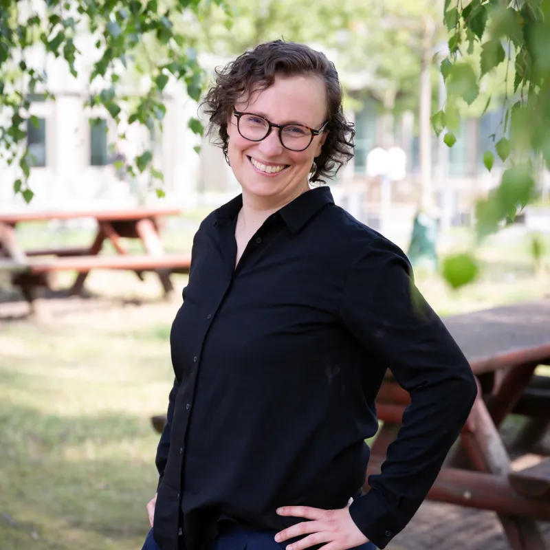 Sabine Rose, Studentin des Masterstudienganges Frühkindliche Bildungsforschung (M.A.) (MAFB) am Fachbereich Sozial- und Bildungswissenschaften der Fachhochschule Potsdam steht in schwarzem Hemd und blauer Hose mit einer Hand in der Hüfte im Freien vor den Bäumen und Holzbänken der Mensa.