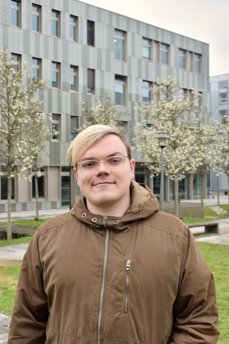 Dominic Bär, ein weißer Mann mit kurzen Haaren und längerem blonden Pony im Gesicht, trägt eine braune Jacke und steht vor einem Gebäude der FHP