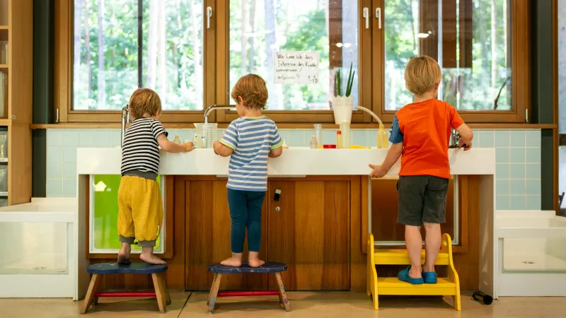 Drei Kinder stehen auf Hockern vor einem großen Waschbecken mit Messbechern