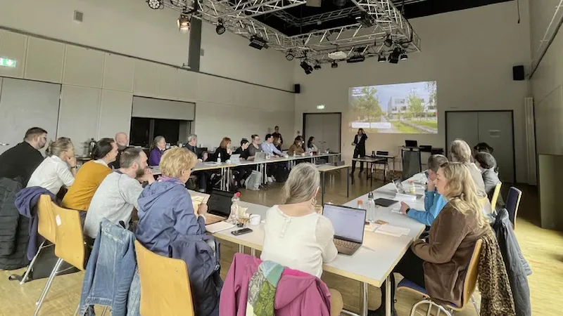 Eine Gruppe Menschen sitzt an Tischen und hört einer vortragenden Person zu