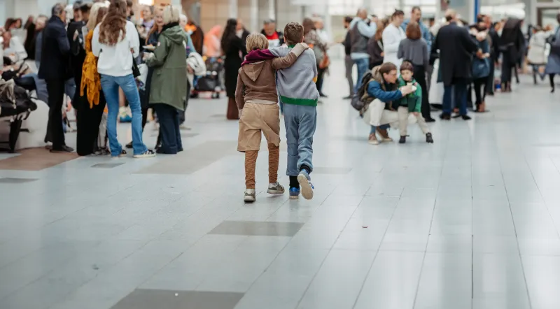 Zwei Kinder gehen Arm in Arm durch eine belebte Halle mit mehreren Menschen im Hintergrund.